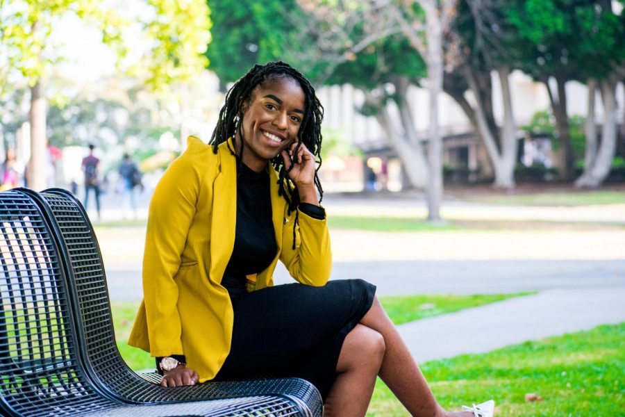 ASI President, Nia Johnson, is smiling and sitting on a campus bench with her mustard jacket, posing with her hand to her cheek