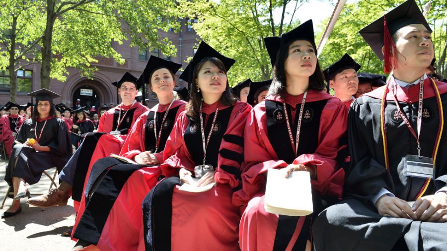 Picture of a bunch of students sitting in a row of chairs with their graduation caps and gowns, smiling as they hear the ceremony