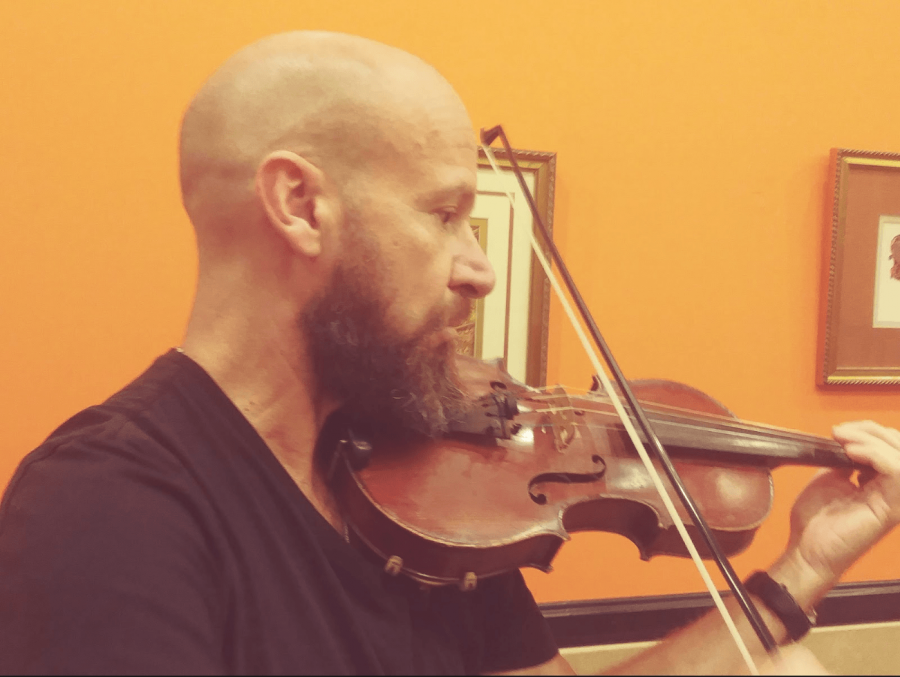 Bald and bearded man is playing the violin inside a music room covered with paintings.