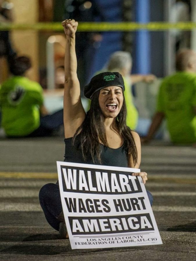 Woman shouting, lifting her fist to the air and is holding a sign titled "Walmart Wages Hurt America"