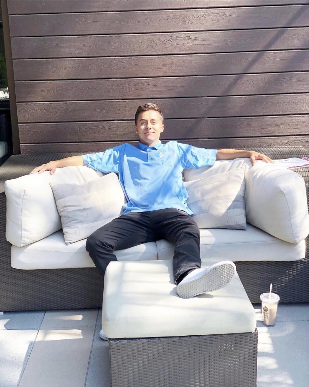 Photo of David Casillas posing on a white outdoor couch with arms on cushions that are on either side and one foot up on an ottoman.