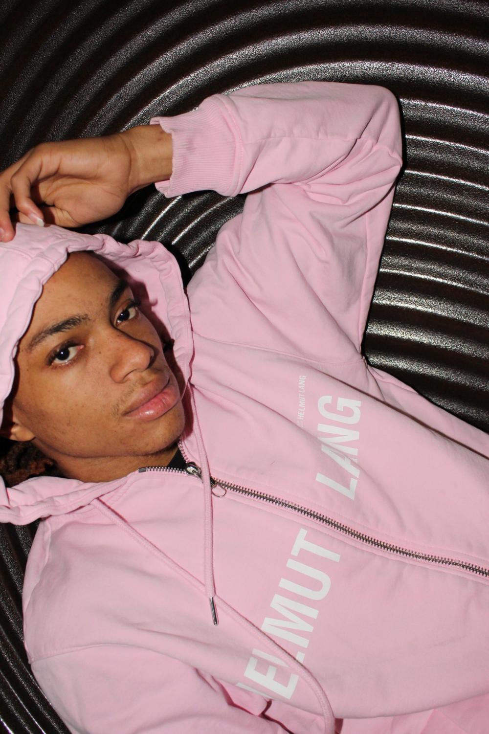 A headshot of Femi Boirard wearing a pink hoodie and posing with his left arm up near his head.
