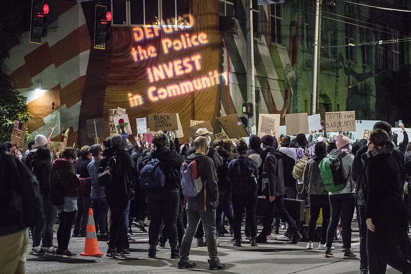 Protestors gathered at night time with text projected on a wall that says, Defund the Police. Invest in Communities.