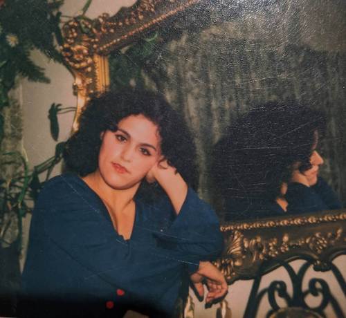 A woman with lipstick and a dark blue dress sits next to a mirror.