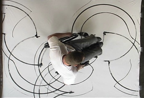 overhead shot of man in wheel chair creating artwork, black half circles on a white canvas