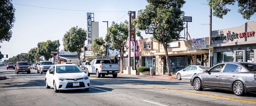 A white car approaching and gray car leaving, driving past single-story beige shops.