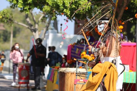 A man dressed up in Día de Los Muertos celebration wear which includes feathers. He is also playing the drum next to an alter set up.