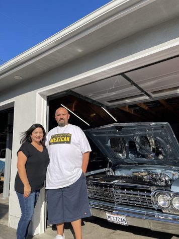 In the photo there are two person looks like couple is standing near the garage and the car bonnet is opened.