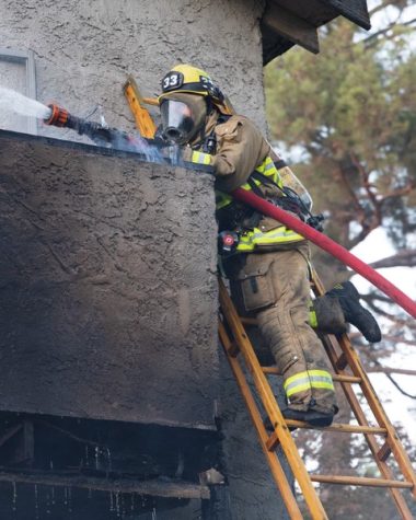 Man in beige and neon yellow firesuit stands on ladder over a balcony and holds red hose with water shooting out