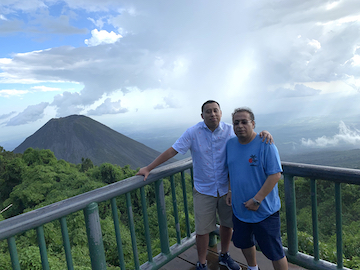 Two men wearing blue shirts stand on a balcony overlooking a view of a dark mountain