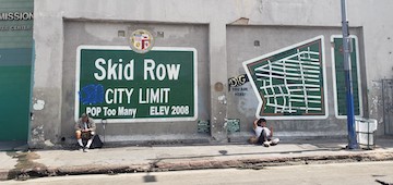 A green sign with white letters reading Skid Row - City limit - too many on a wall in front of two men sitting on sidewalk