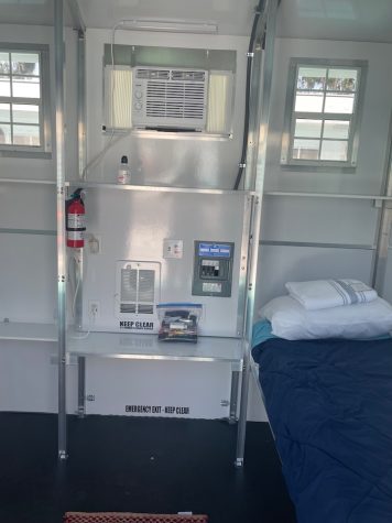 Inside a small room with metallic bars help form shelves and a bed with white pillowcase and dark covers