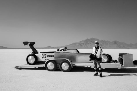 Black and white photo of a person standing in front of an old looking car in the middle of the desert. 