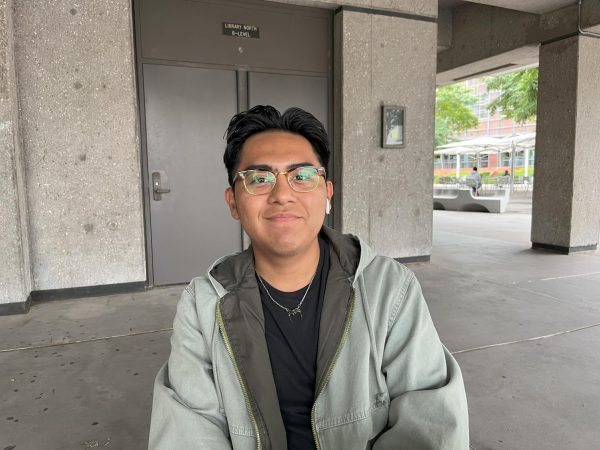 A photo of a college student with yellow tinted glasses, a black shirt, and a seafoam green jacket on. He also has an airpod in his left ear.