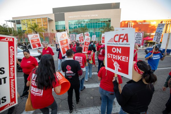 A group of people wearing red and black shirt cross a street while holding signs that say "CFA ON STRIKE!"