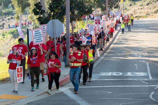 A group of people holding signs and dressed in red shirts and traffic bests walk along the sidewalk.
