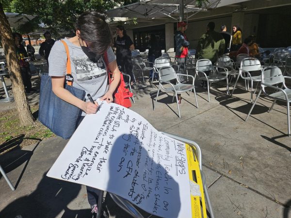 A person wearing a gray shirt, holding a marker and writing on a poster board, while standing on cement outside. Other people standing behind person with poster board.