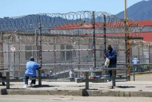 Two men wearing blue in a yard with a fence and barbed wire on top