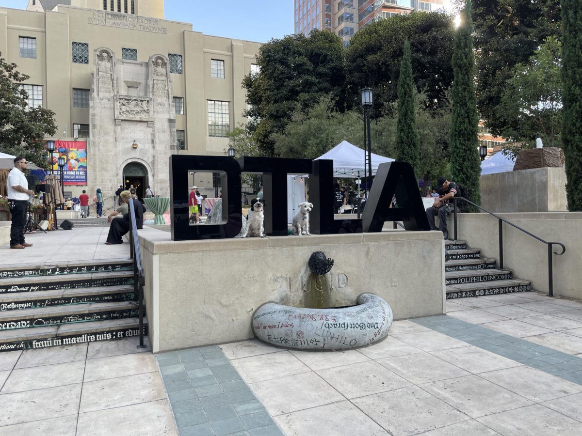 Front view of Dog Day LA 2025 gathering in Maguire Gardens at LA Central Library. Dogs Gonchi (left) and Archie (right) pose under a DTLA sign on Sept 12 2025.